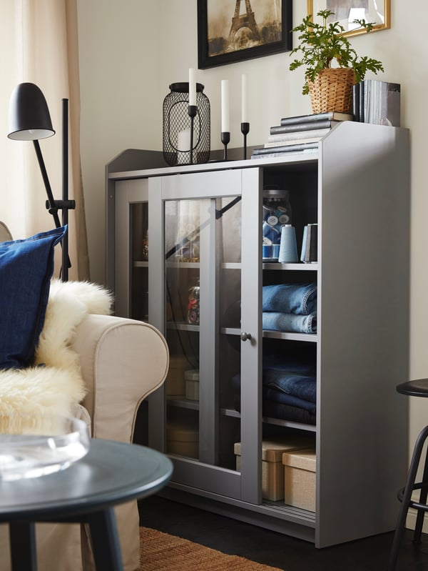 Des jeans pliés et des boîtes sont rangés dans une vitrine. Elle côtoie un canapé beige clair et un tabouret noir.