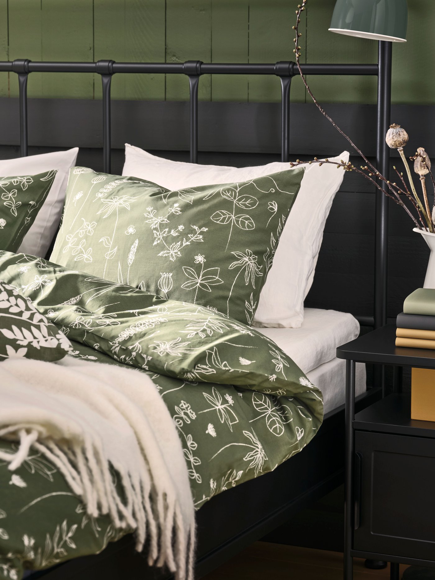 Un dormitorio con una cama de metal negra con ropa de cama verde. Al costado de la cama un velador negro.
