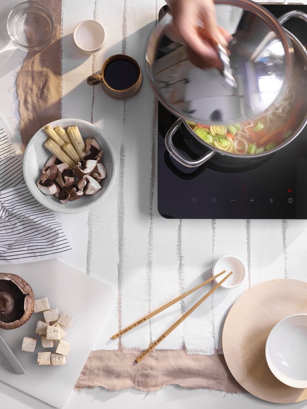 A hand lifts the lid off a pot of vegetables on a black portable hob placed on a white table with cups and chopsticks.