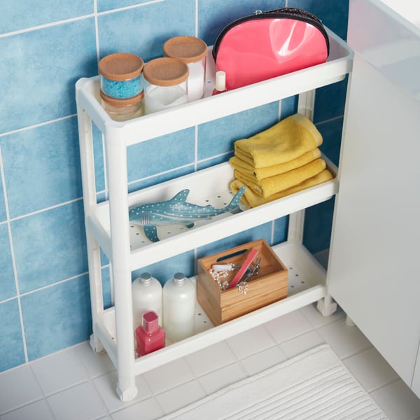 A white VESKEN trolley against a blue-tiled wall has glass jars with lids, small yellow towels and a toy shark stored on it.