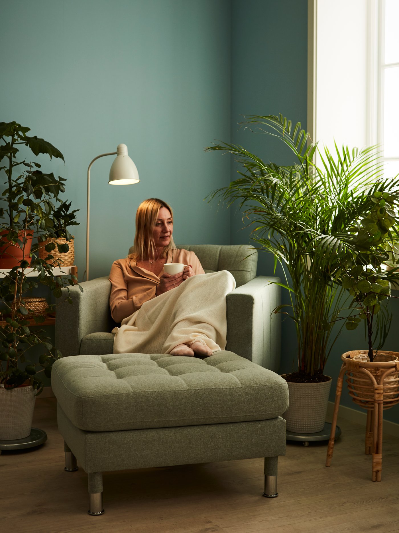 Living room with green walls, woman sitting on an armchair with her legs resting on a footstool, floor lamp, and plant stand.