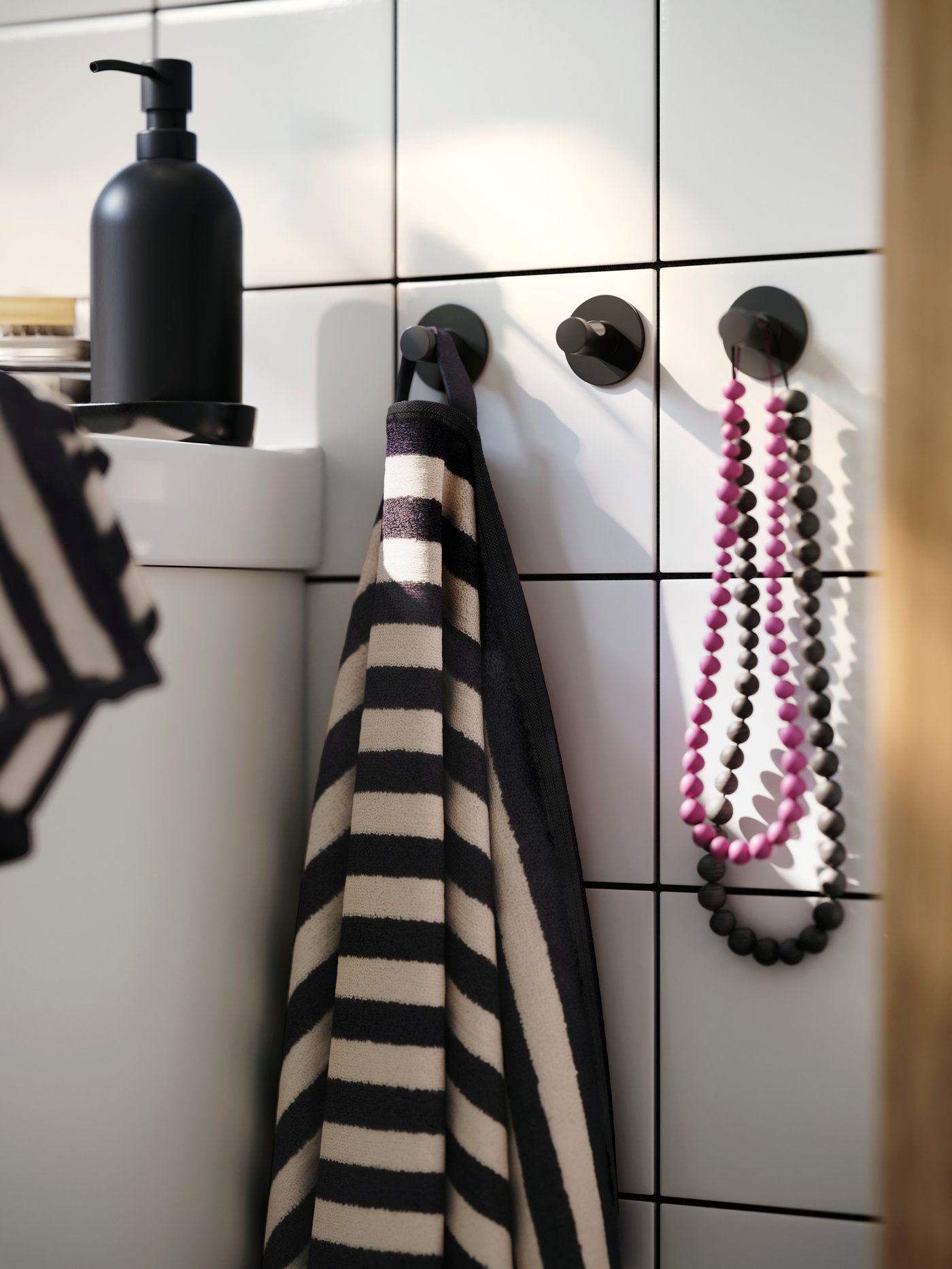 Three black SKOGHALL towel racks hang in a row against white bathroom tiles. Black and white striped towels and a necklace.