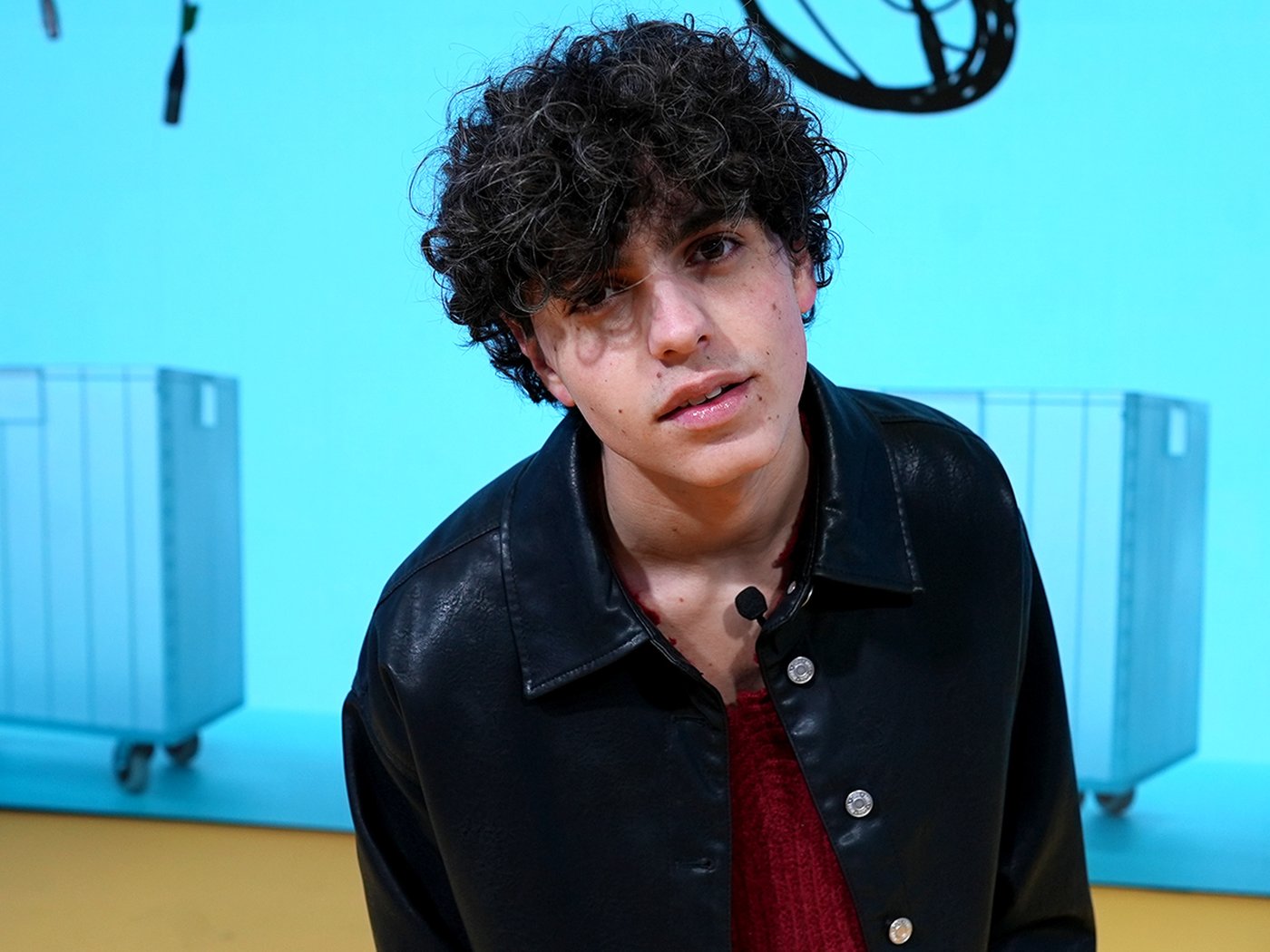 Niveo standing on stage in a black shirt and necklace with a blue colour with IKEA products as a background that comes from the digital display used on stage during Milan design week 2024 at the IKEA venue.