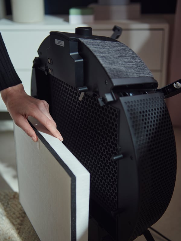 A woman removes the particle filter from the front of a black floor model version of the STARKVIND air purifier.