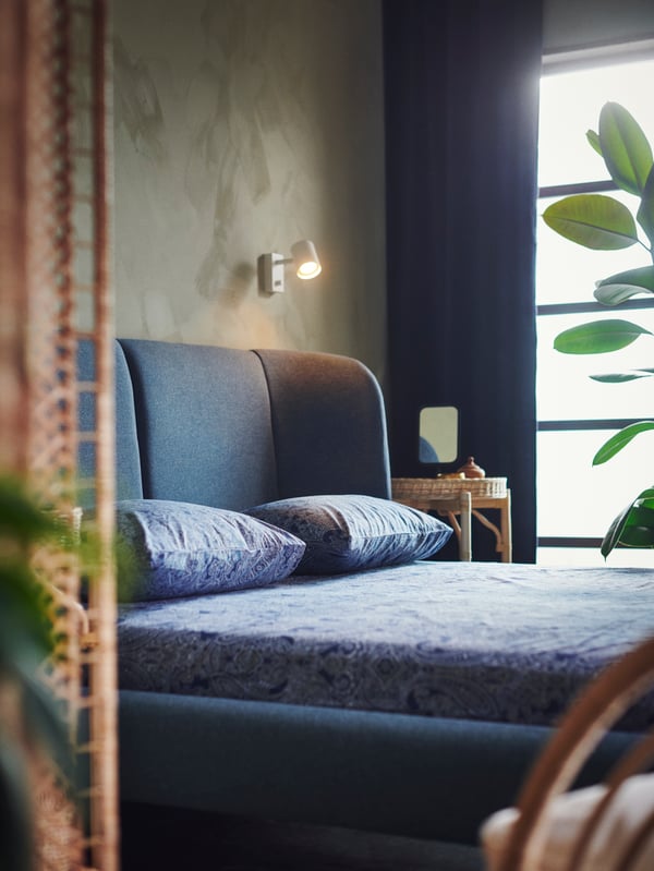 سرير يتنجيد TUFJORD أزرق مغطى ببياضات سرير JÄTTEVALLMO. ومصباح توجيهي مثبت في الحائط.