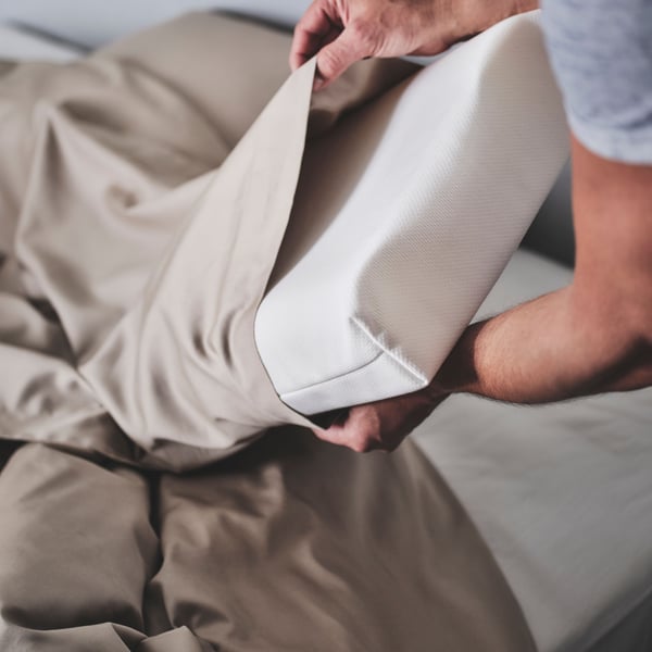 Personne glissant un oreiller ergonomique côté/dos PAPEGOJBUSKE blanc dans une taie d’oreiller en satin beige.