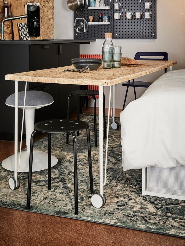 A wooden tabletop on white legs with castors, between a bed, two black stools and a white/grey NILSERIK standing support.