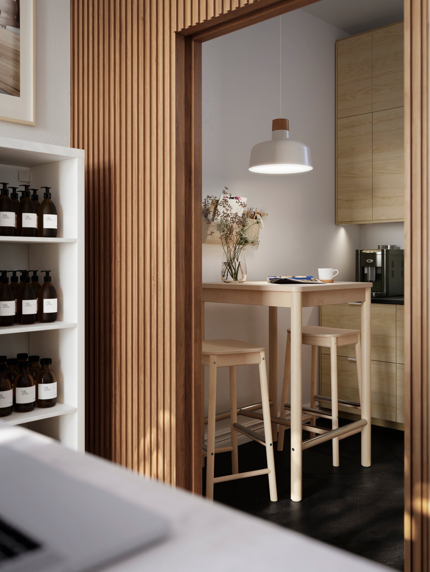 A slatted-wood wall opening into a backroom with RÖNNINGE birch bar stools and table, flowers in a vase and a coffee machine.