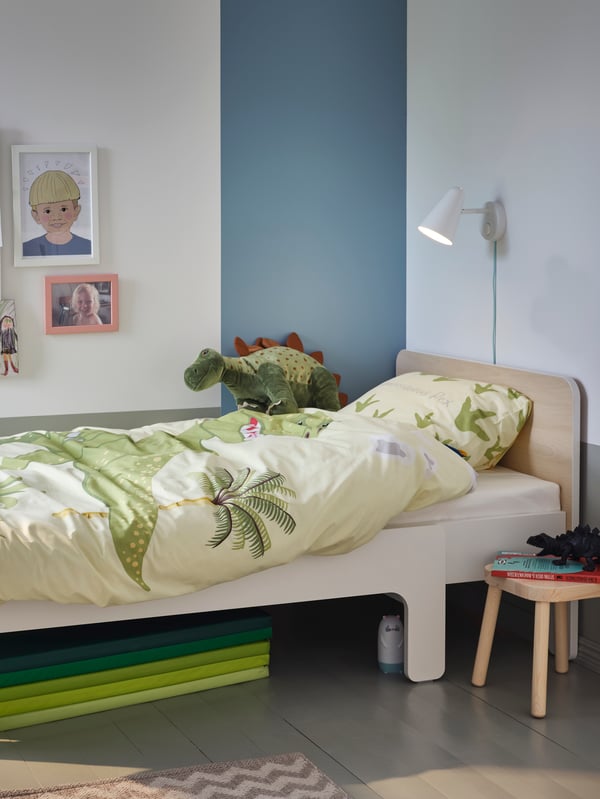 Una cama extensible SLÄKT con un cobertor JÄTTELIK de tonos verde claro en una habitación infantil con piso gris oscuro y muros blancos. Bajo la cama se ven unas colchonetas PLUFSIG y a un costado, a modo de velador, un piso de madera FLISAT. 