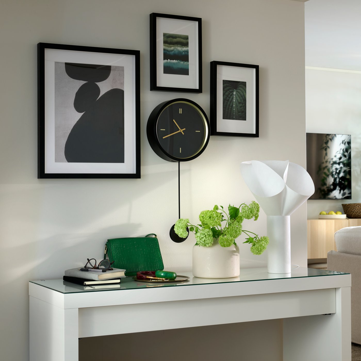 Framed art hanging around a black STURSK wall clock, all placed above a console table with a white BÄCKNATE table lamp on it.
