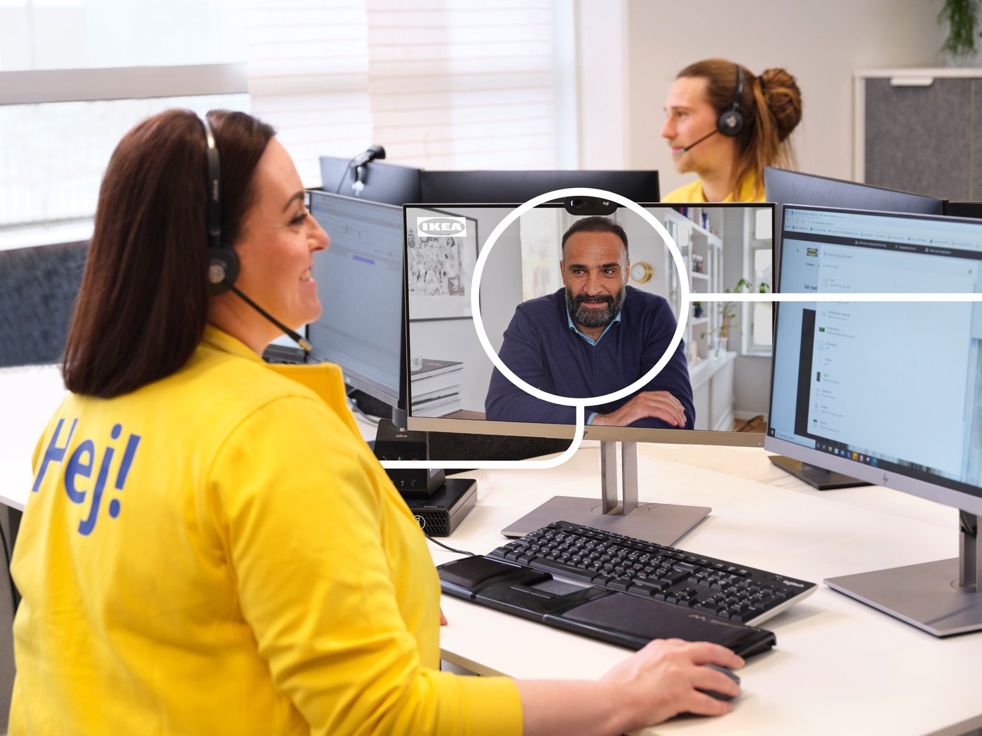 Person arbetar vid en datorarbetsplats med headset och genomför ett videosamtal på en skärm. Flera bildskärmar, tangentbord och kontorsutrustning syns i en kundservice- och kontorsmiljö.
