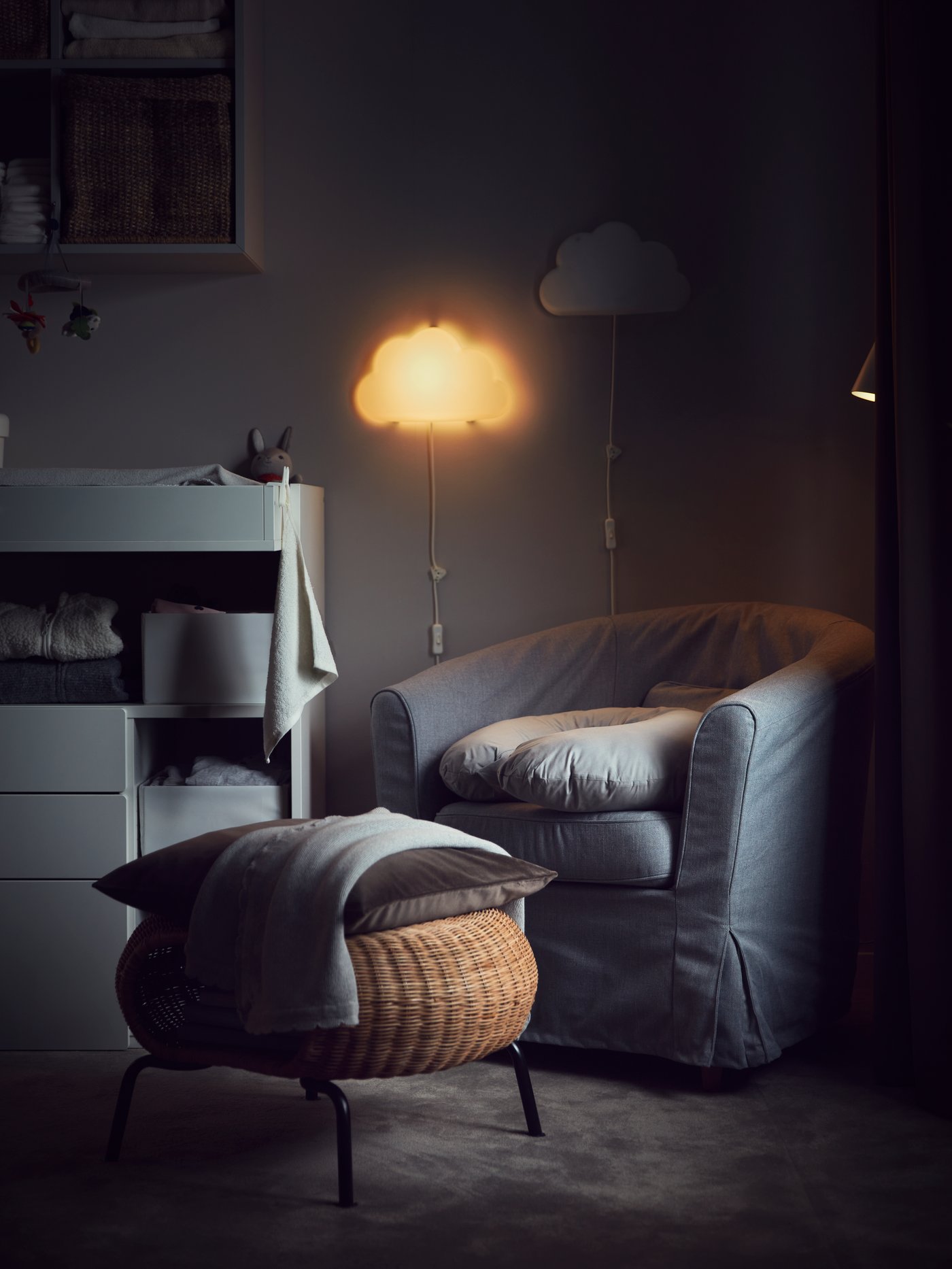 Mysigt, svagt upplyst rum med en grå fåtölj som har en kudde och filt. Framför fåtöljen står en korg i rotting med ytterligare en kudde och filt ovanpå. Till vänster finns ett vitt skötbord med hyllor fyllda med vikta kläder och andra föremål. Ovanför skötbordet hänger en molnformad lampa som sprider ett varmt sken, samt en annan molndekoration.