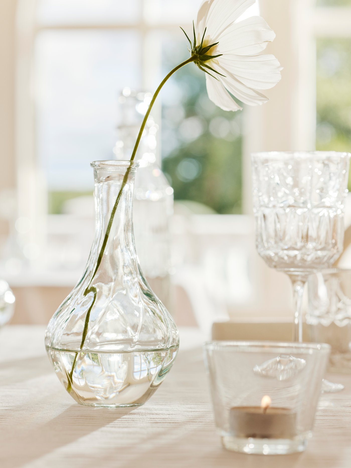 A VILJESTARK clear vase with a single white flower, a GALEJ tealight holder and a SÄLLSKAPLIG wine glass are on a table.