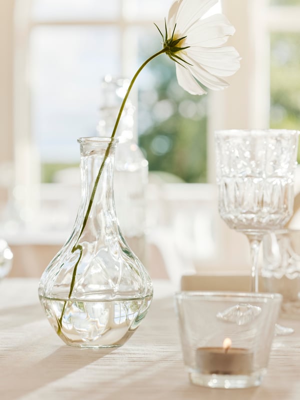 Sur une table, se trouvent un photophore GALEJ, un verre à vin SÄLLSKAPLIG et un vase clair VILJESTARK dans lequel est disposée une seule fleur blanche.