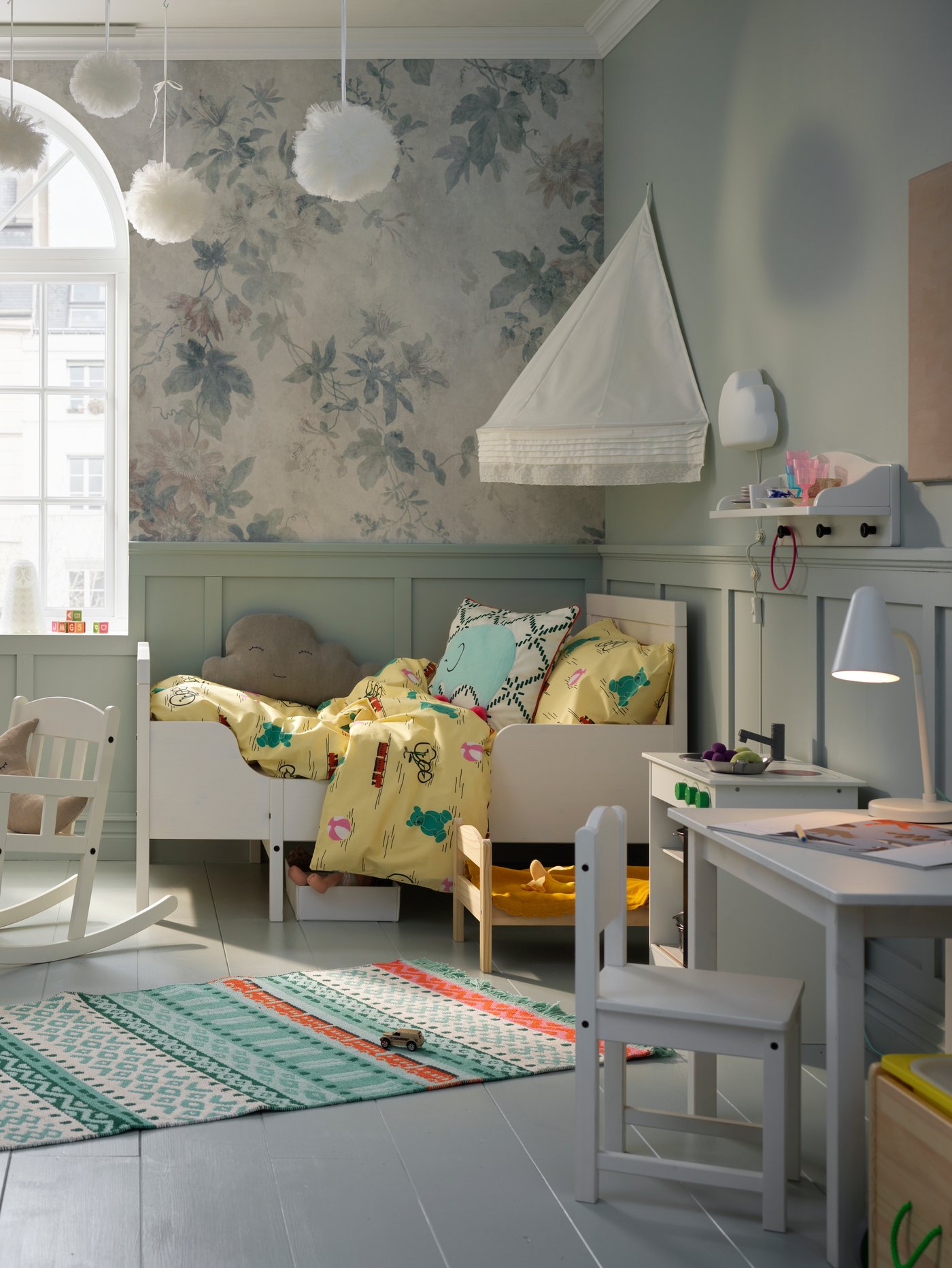 A children's room featuring a white extendable bed, a quilt cover, a bed canopy a rocking chair, a play kitchen and a rug.