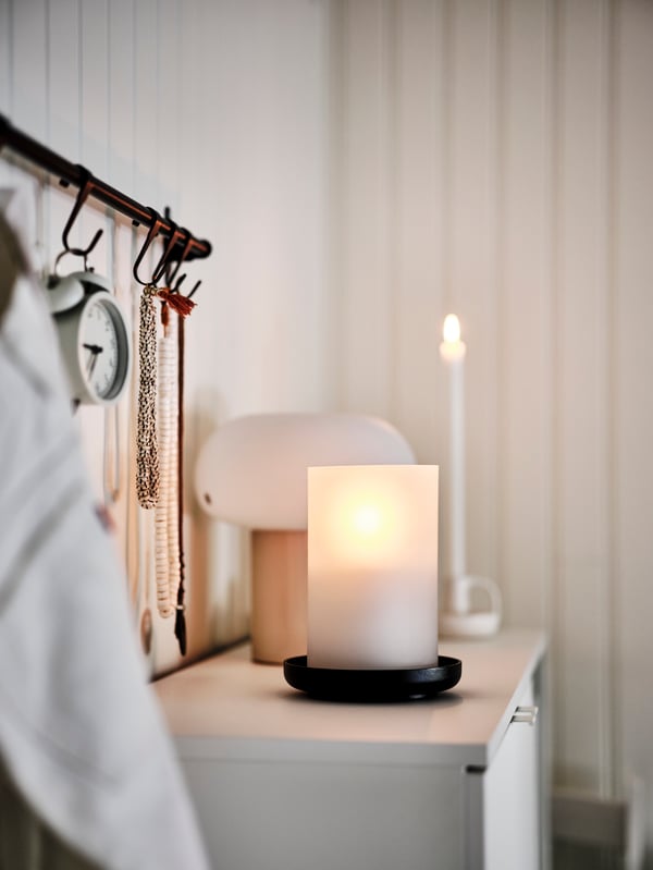 A black HULTARP rail with hooks above a lit candle inside a frosted glass/black HEDERVÄRD lantern next to a DEJSA table lamp.