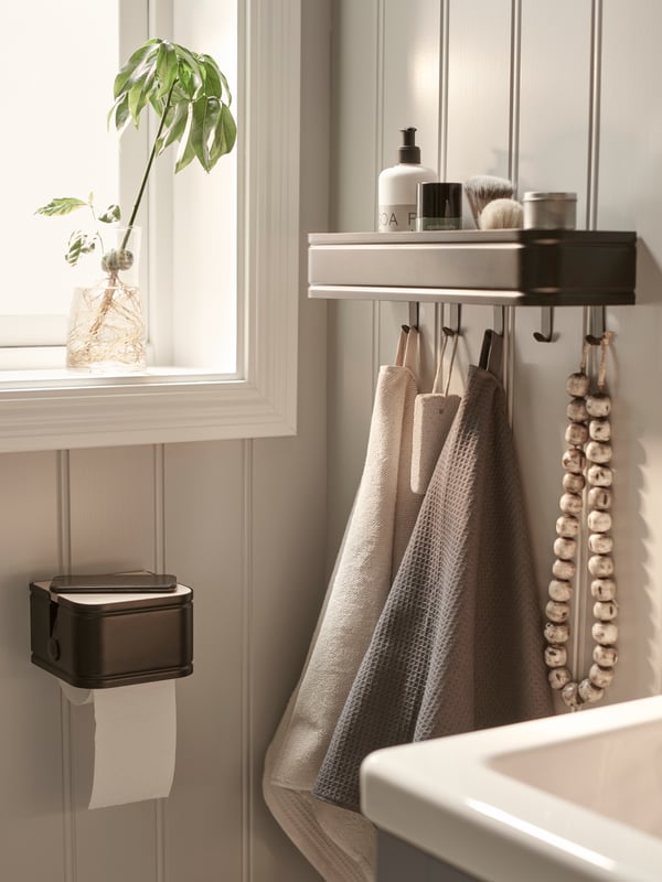 A LILLASJÖN toilet roll holder with a mobile phone on top perpendicular to a LILLASJÖN shelf with hooks on a panelled wall.