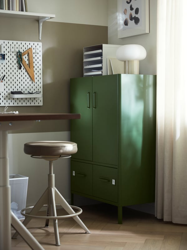 Un meuble IDASEN vert est placé dans le coin d’un espace de travail, à côté d’un bureau et d’un tabouret taupe. 