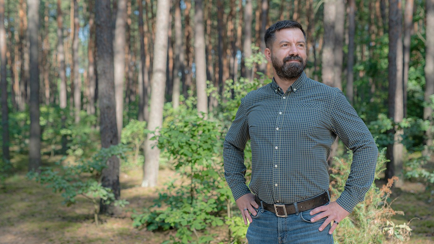 Maciej stoji u osunčanoj šumi i nosi kariranu košulju i farmerke.