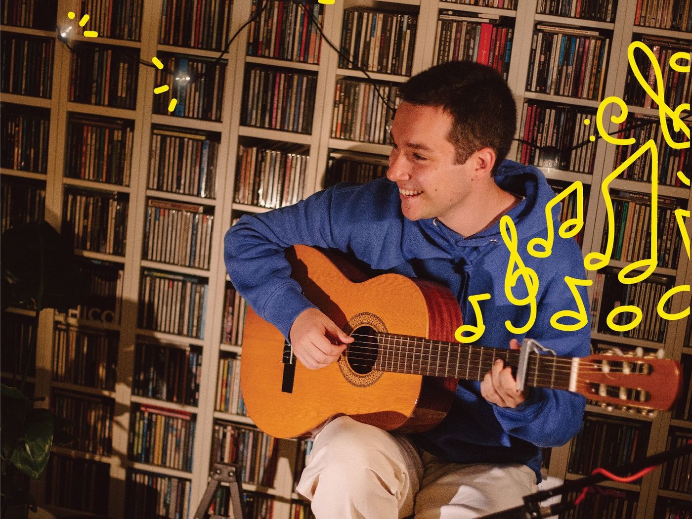 A man playing the guitar in front of a bookcase wall decorated with string lights.