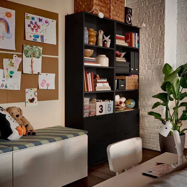 A black BRIMNES bookcase filled with books and decorative boxes, beside a cosy reading nook with soft-toy animals.