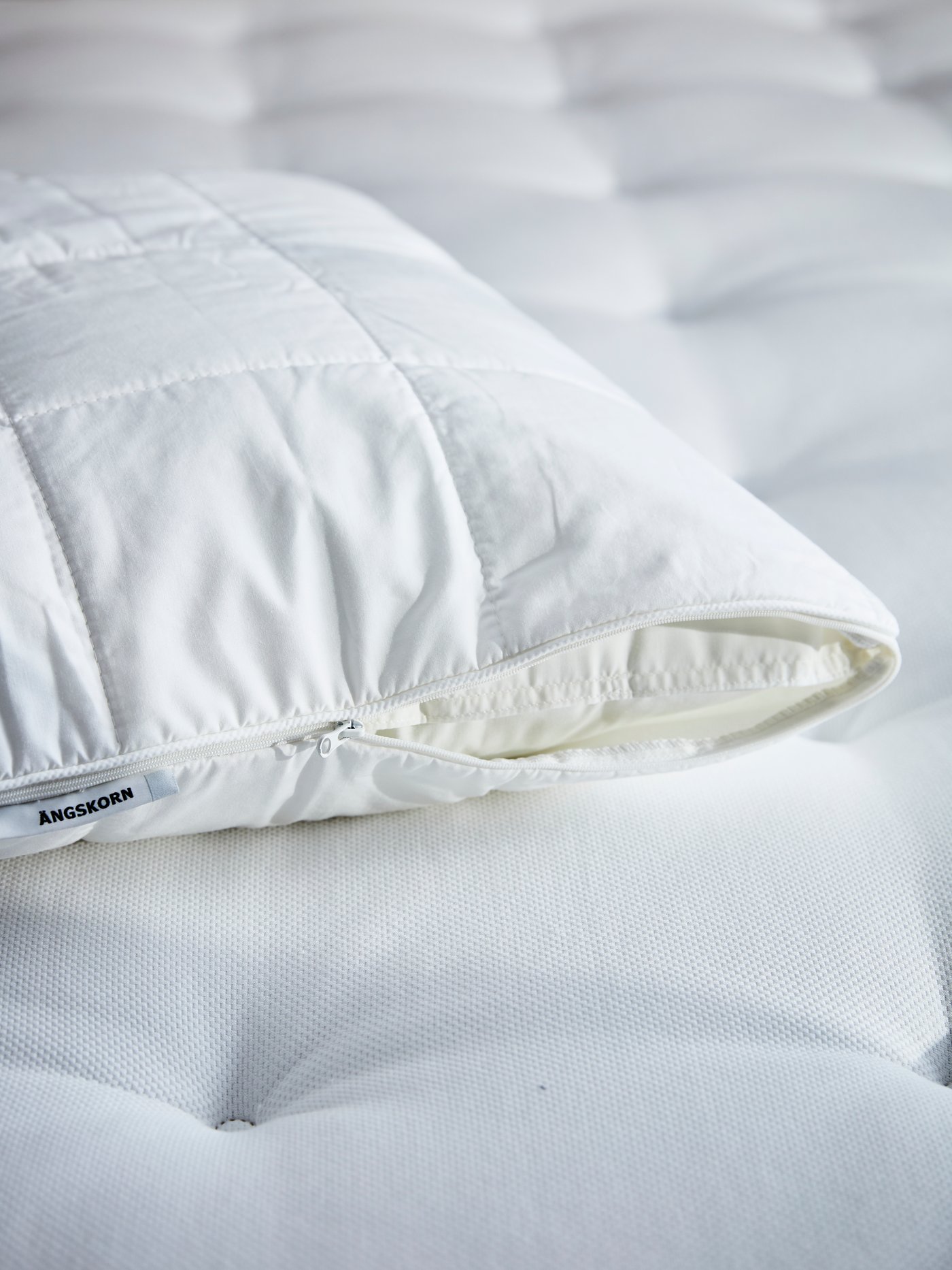 Bantal di dalam pelindung bantal ÄNGSKORN yang dizip separuh diletakkan di atas tilam berwarna putih.