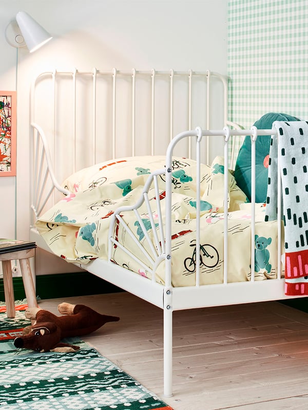 Una cama extensible MINNEN blanca con ropa infantil colorida. Sobre el cabecero de la cama una lámpara infantil.