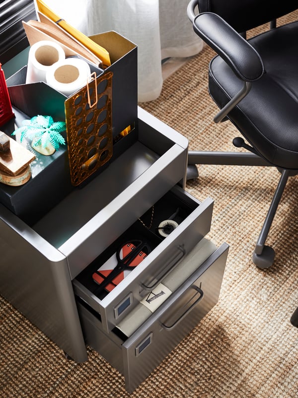 A black chair beside a storage unit, drawers open, holding files and items with various office supplies on the top.