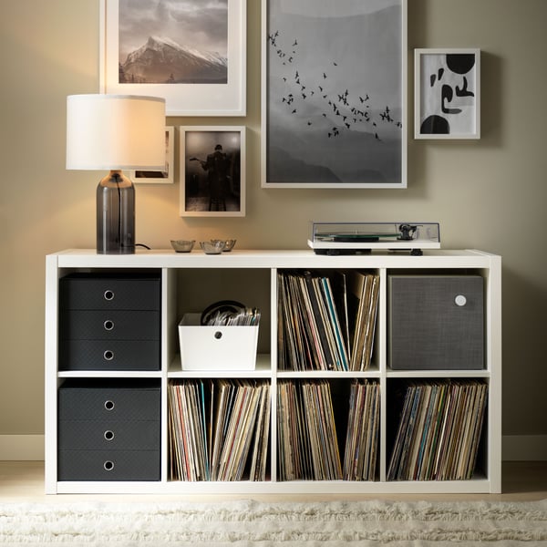 Un estante KALLAX de ocho compartimientos, tres de ellos con cajas organizadoras, el resto con vinilos. Sobre el estante hay una lámpara y en la pared cuadros decorativos.