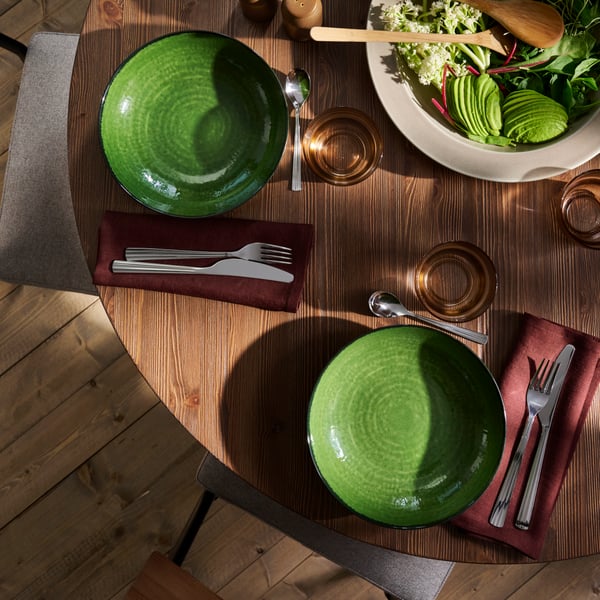 A wooden dining table with PELARKAKTUS side plates in green, set alongside cutlery, napkins, glasses and salad bowl.