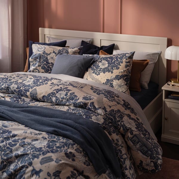 A pink bedroom with a white bed made with PJÄXE bed linen in a floral pattern. Next to the bed stands a white bedside table.
