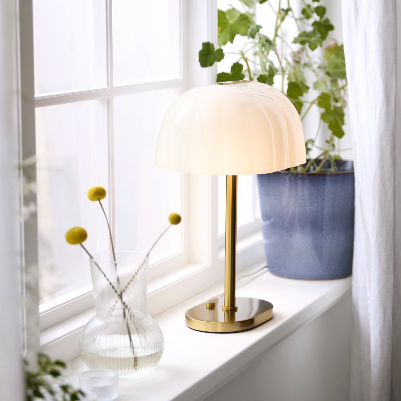 Blå kruka i fönster med vit gardin och lampa med mässingsfot.
