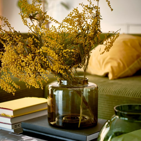 A brown KONSTFULL vase with yellow branches in it, in front of a green sofa.