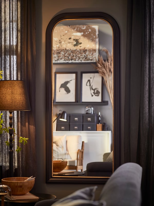 A black ALMARÖD mirror hangs in a living room, reflecting picture ledges with framed artworks on them on the opposite wall.