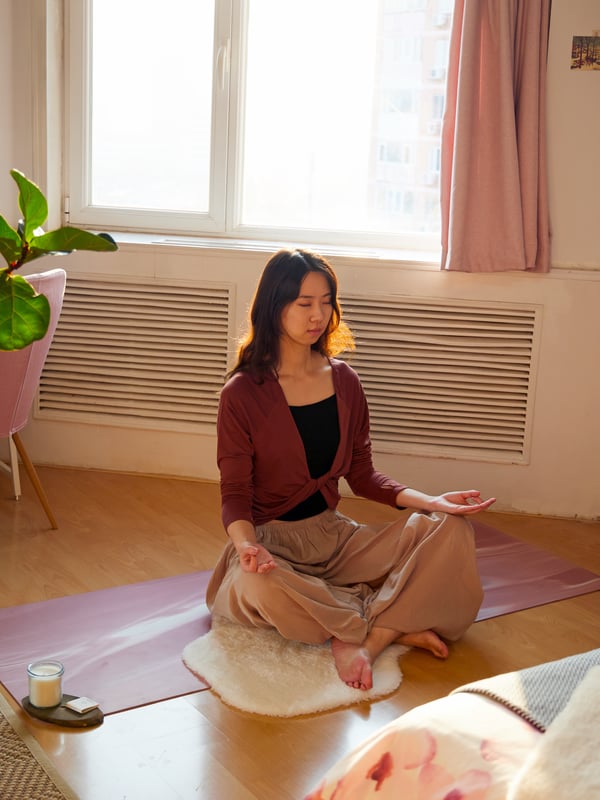 Žena meditira na podu u svojoj sobi pored prozora, a kao oslonac koristi mekani beli TOFTLUND tepih.
