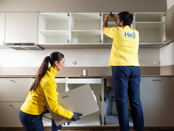 Dva delavca, oblečena v rumene uniforme IKEA, sestavljata ali nameščata kuhinjske omare. Eden pritrjuje zgornjo omarico, drugi pa drži vrata spodnje omarice. To prikazuje storitve podjetja IKEA, kot je montaža kuhinj.