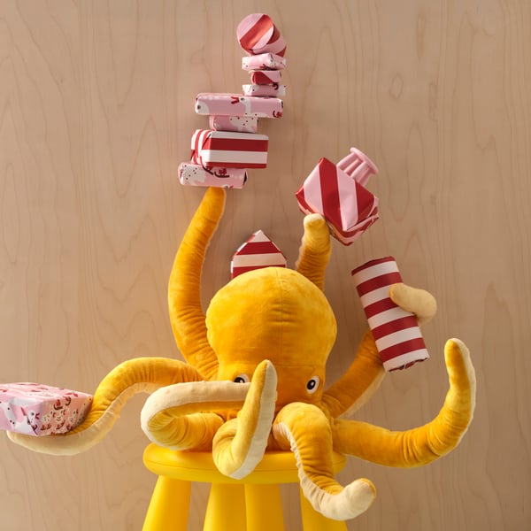 A yellow BLÅVINGAD soft toy octopus is sitting on a MAMMUT stool, holding several wrapped gifts in five of its eight arms.