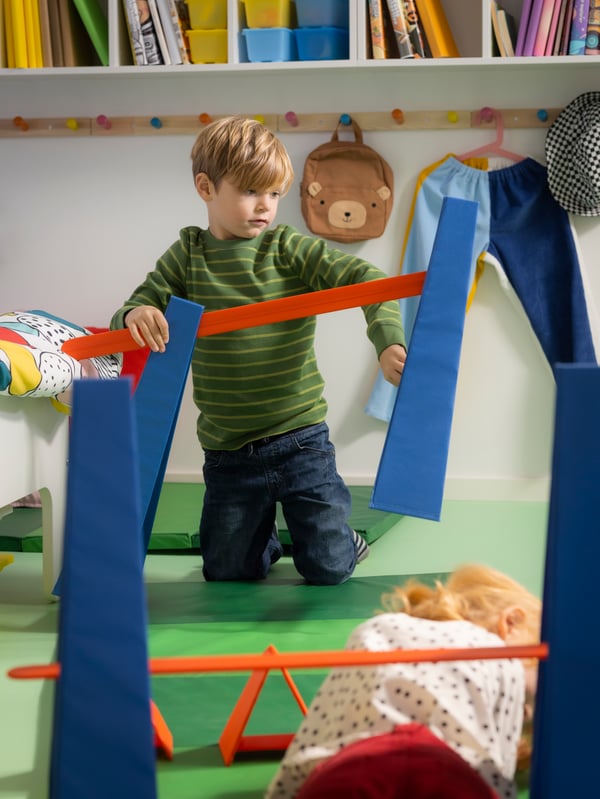 Twee kinderen spelen met een SKRATTMÅS hindernisbaan in een ruimte met twee groene, opvouwbare PLUFSIG gymnastiekmatten op de grond.