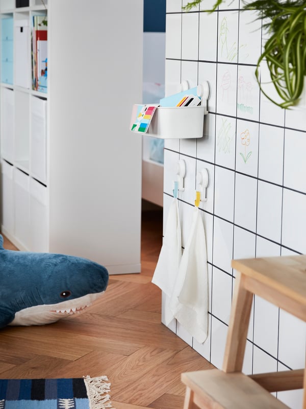 A white tiled wall with TISKEN basket and hooks with suction cups attached to it, they hold children’s toys and small towels.