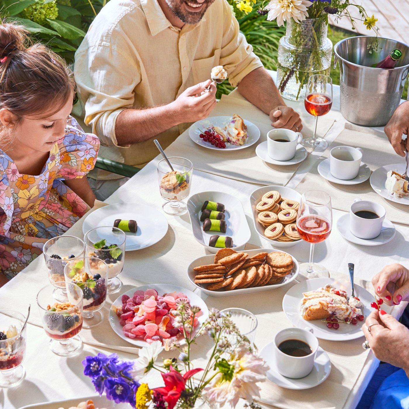 En familie fejrer svensk midsommer udenfor og er samlet om spisebordet med mange snacks, mad og kaffe.