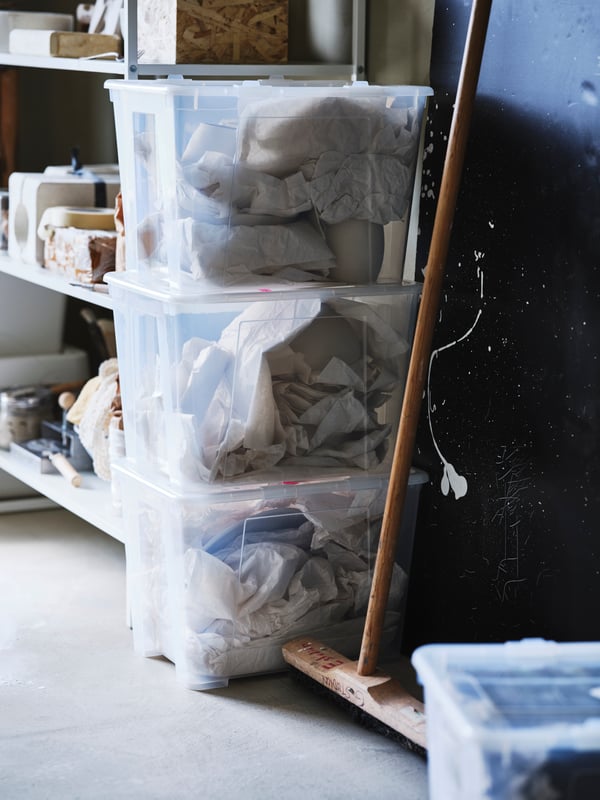 A softly sunlit room with assorted storage on shelves and in a stack of SAMLA storage boxes. Next to it stands a broom.