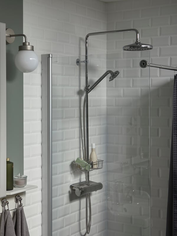 A white-tiled shower with a glass door, a chrome-plated VOXNAN head shower and shelf holding two shower products.