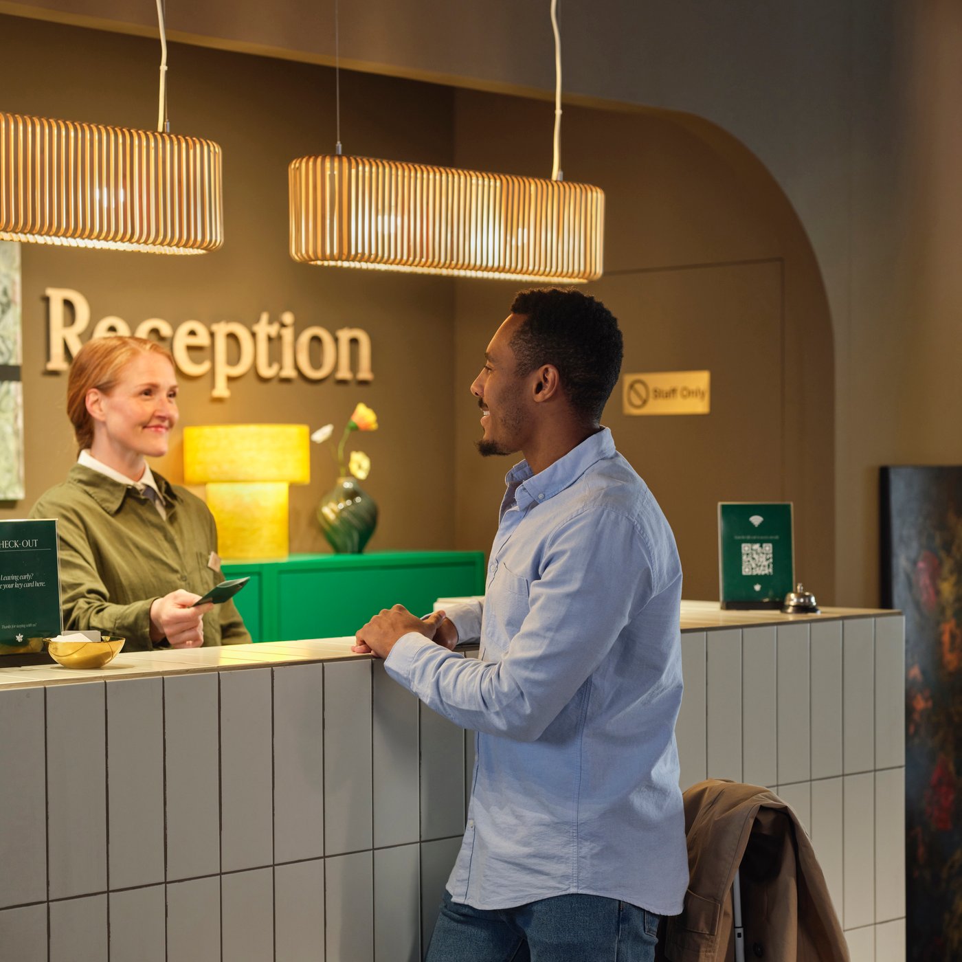 Hotel reception desk; staff hands card to guest under warm pendant lights, “Reception” sign visible behind counter.