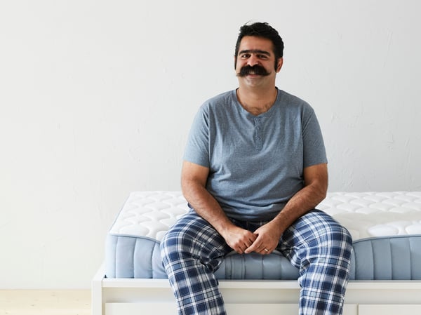 A man with a moustache and wearing a greyish-blue t-shirt and blue plaid pyjama pants sits on the edge of a mattress.