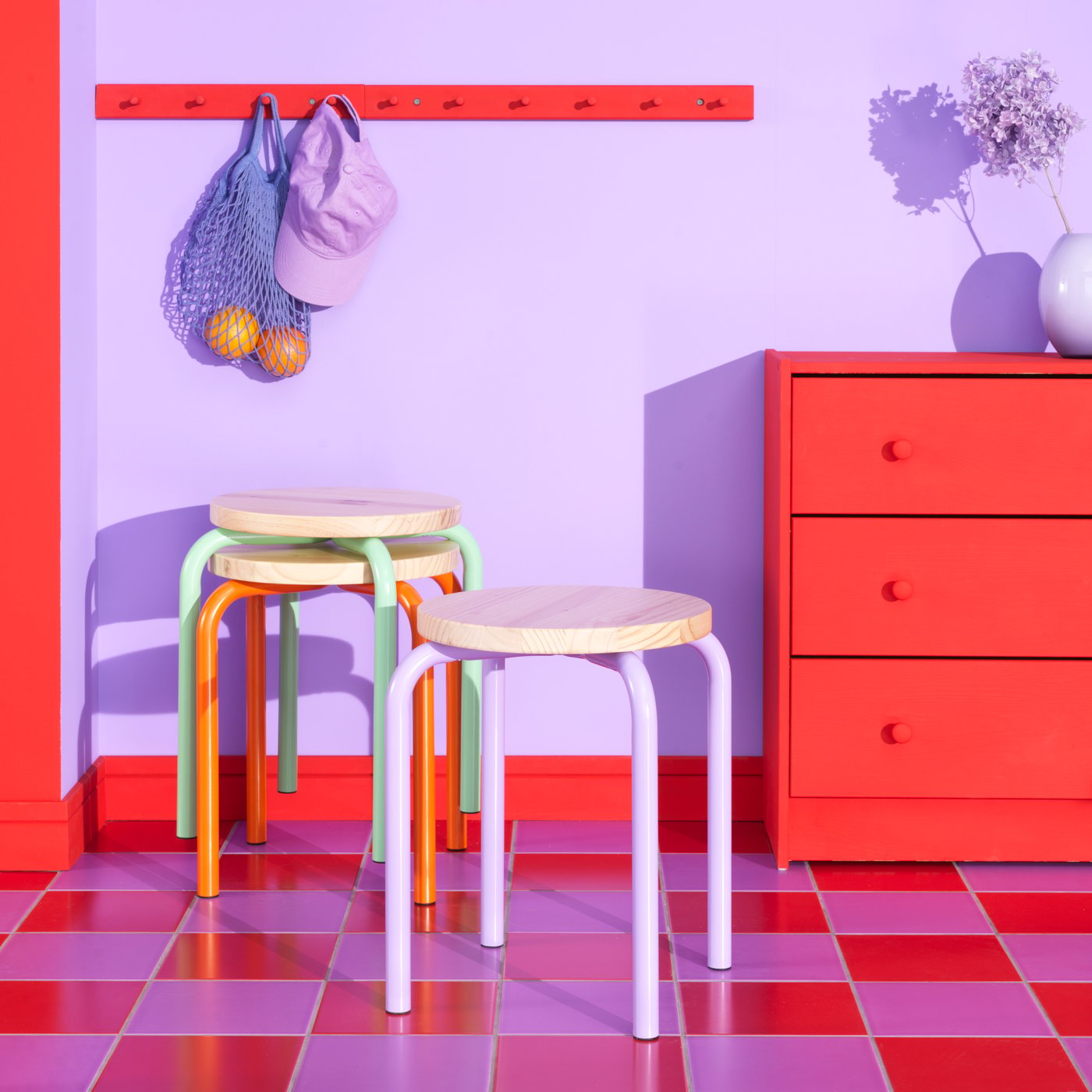 Ruimte in paars en rood met een geruite vloer in dezelfde kleur en IKEA krukjes, ladekast en kleerhangers.
