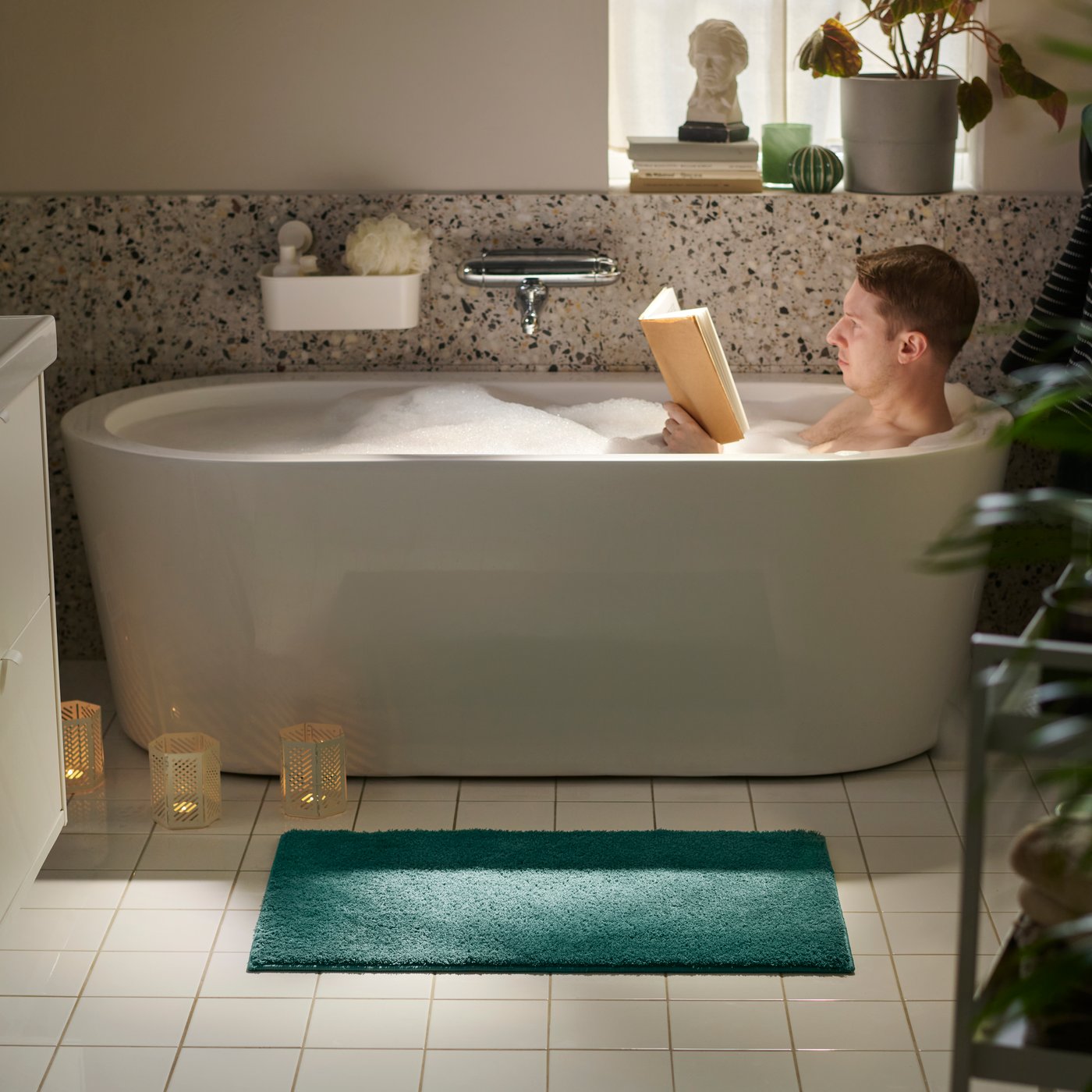 A bathroom showing a BACKMÅRA bath mat on the floor beside a bath tub, where a person relaxes in the bath.