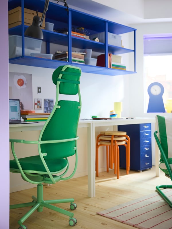Ein hellgrüner LÖPARBANA Gaming-Stuhl neben einem Schreibtisch mit blauen Regalen an der Wand dahinter. Blaue und orangefarbene Details im Hintergrund.