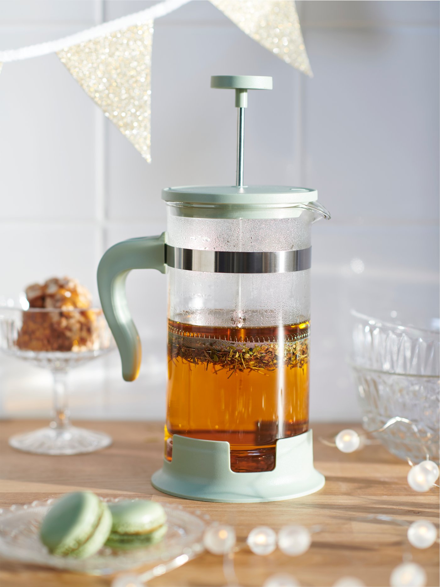 An UPPHETTA coffee/tea maker half-full of tea on a wood surface with a SÄLLSKAPLIG plate and bowls with confectionaries.
