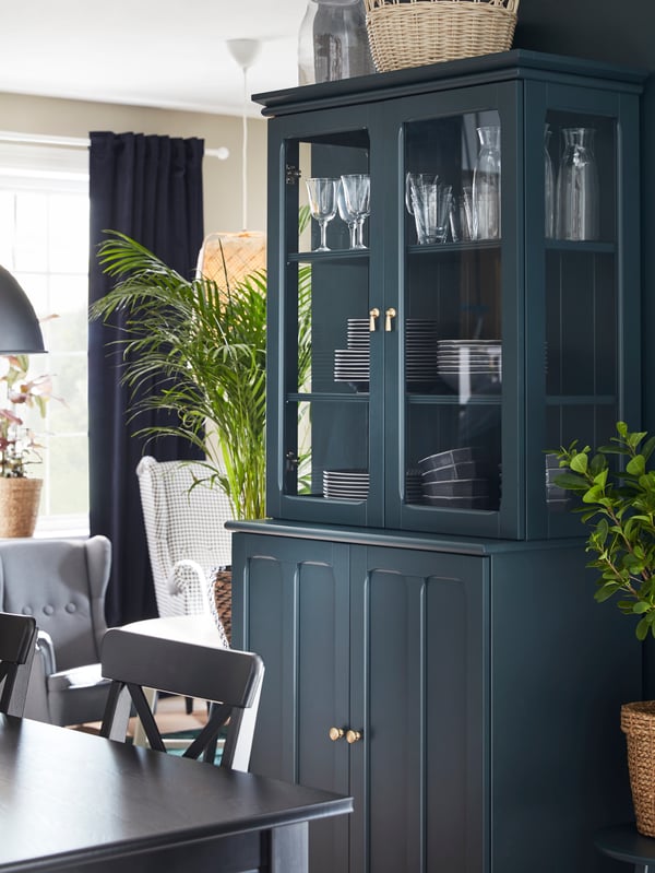 A dark blue-green LOMMARP cabinet with glass doors stands next to a dining table, dinnerware stands behind the glass doors.