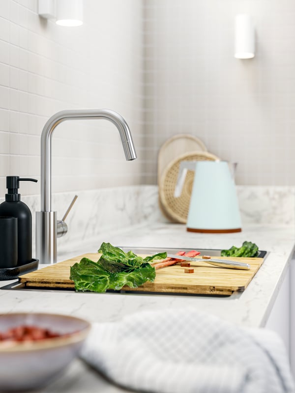 A kitchen countertop with a NORRSJÖN chopping board, fresh vegetables, and a kettle, all set against a tiled backsplash.